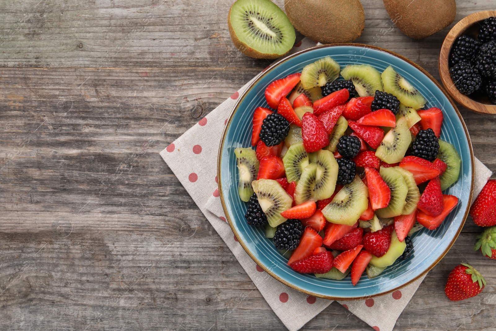 Photo of Plate of delicious fresh fruit salad and ingredients on wooden table, flat lay. Space for text