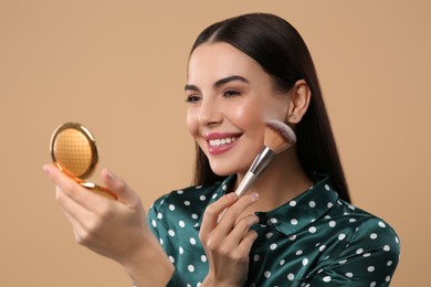 Happy woman with cosmetic pocket mirror applying makeup on light brown background
