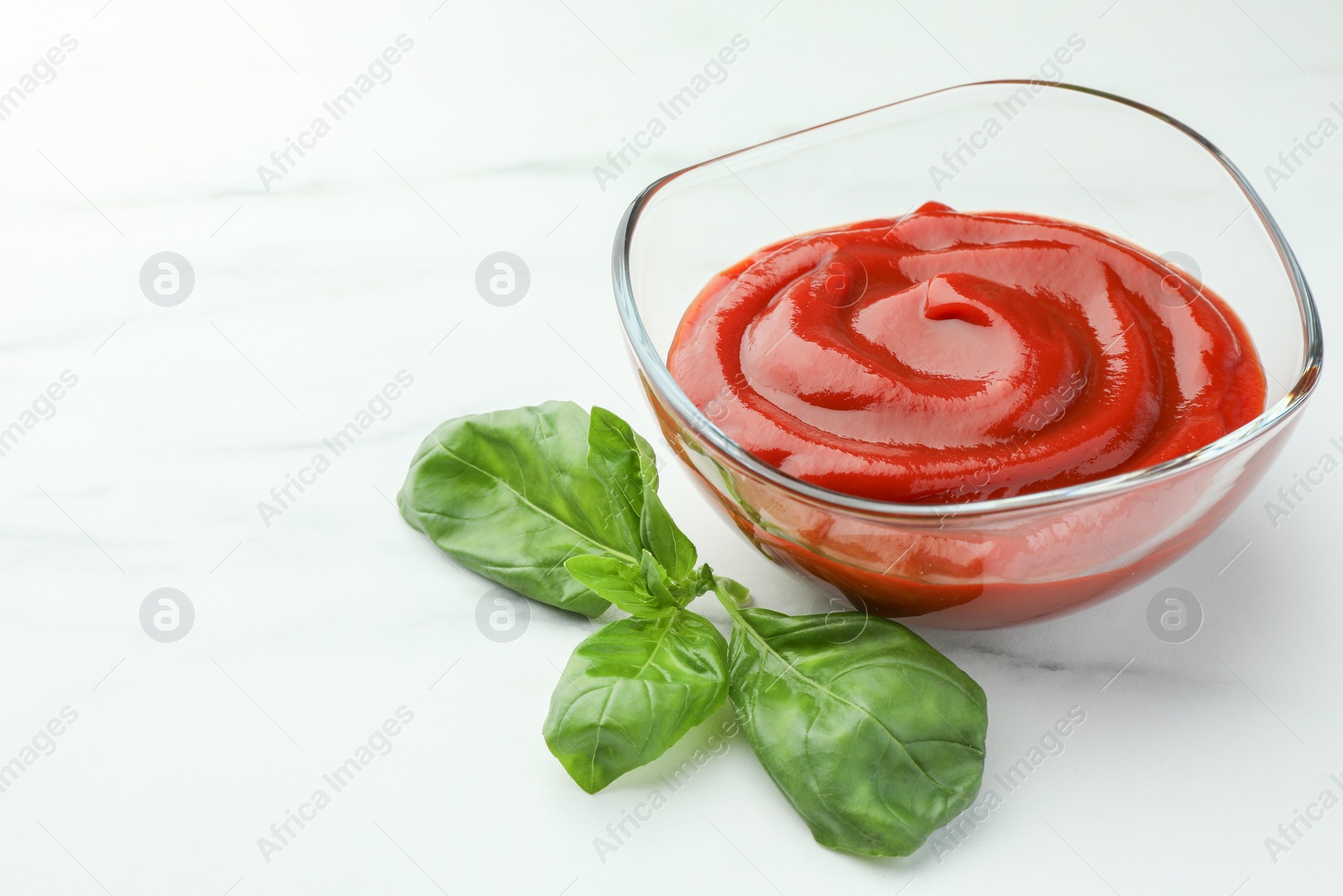 Photo of Bowl of tasty ketchup and basil on white marble table, closeup. Space for text