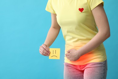 Image of Woman holding sticky note with drawn sad face and suffering from cystitis symptoms on light blue background, closeup