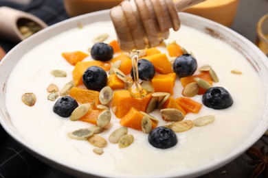 Photo of Pouring honey onto delicious semolina pudding with blueberries, pumpkin and seeds, closeup