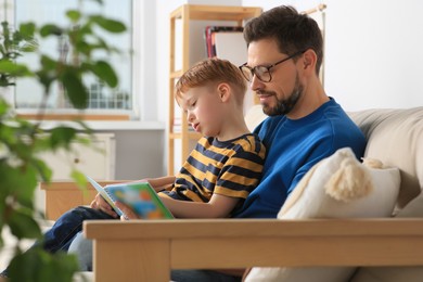 Photo of Father reading book with his son on sofa in living room at home