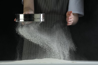 Woman sieving flour at table against black background, closeup