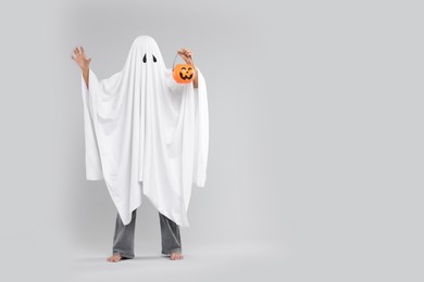 Woman in white ghost costume holding pumpkin bucket on light grey background, space for text. Halloween celebration