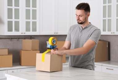 Photo of Man taping box with adhesive tape dispenser in kitchen, space for text