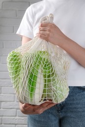 Photo of Woman holding string bag with fresh Chinese cabbages near white brick wall, closeup