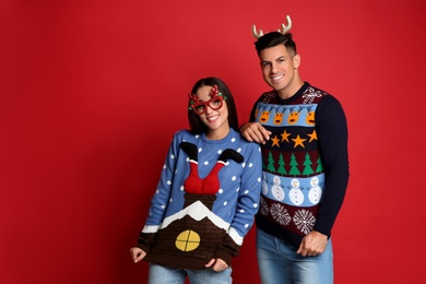 Photo of Couple in Christmas sweaters, deer headband and party glasses on red background, space for text