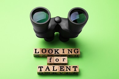 Photo of Staff recruitment concept. Phrase Looking For Talent made of wooden cubes and binoculars on light green background, above view