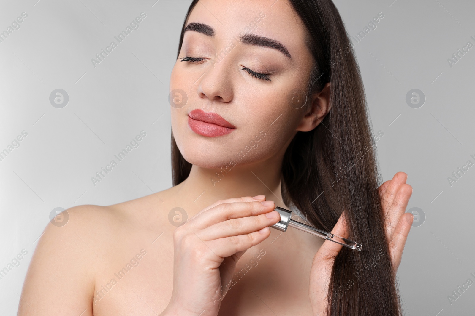 Photo of Young woman applying essential oil onto hair on light grey background