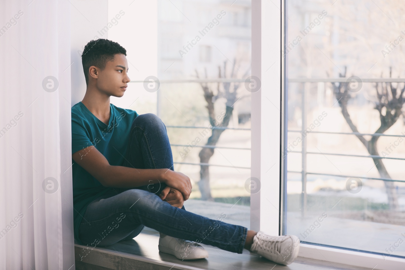 Photo of Upset African-American teenage boy sitting alone near window