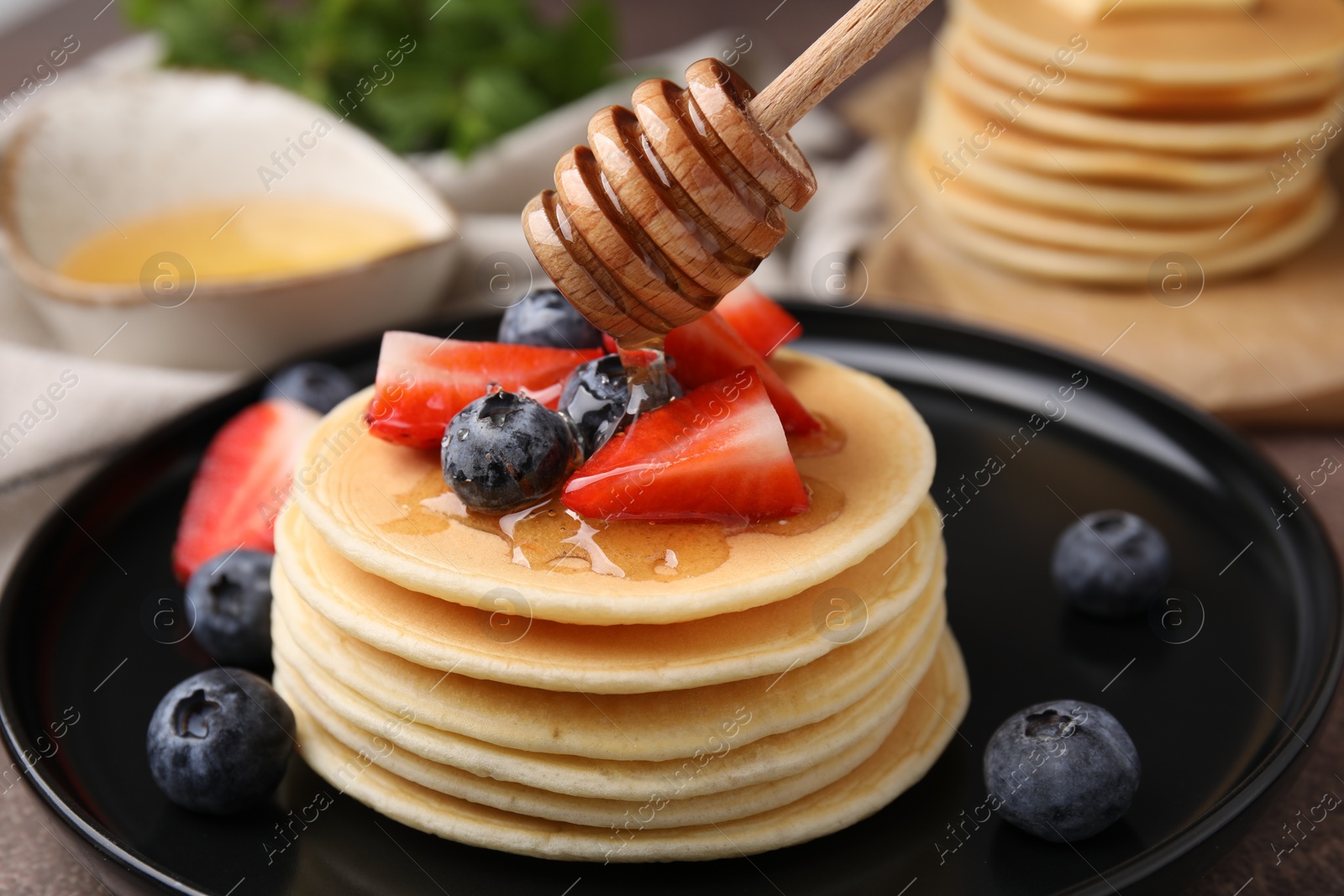 Photo of Pouring honey from dipper onto delicious pancakes with strawberries and blueberries at table, closeup