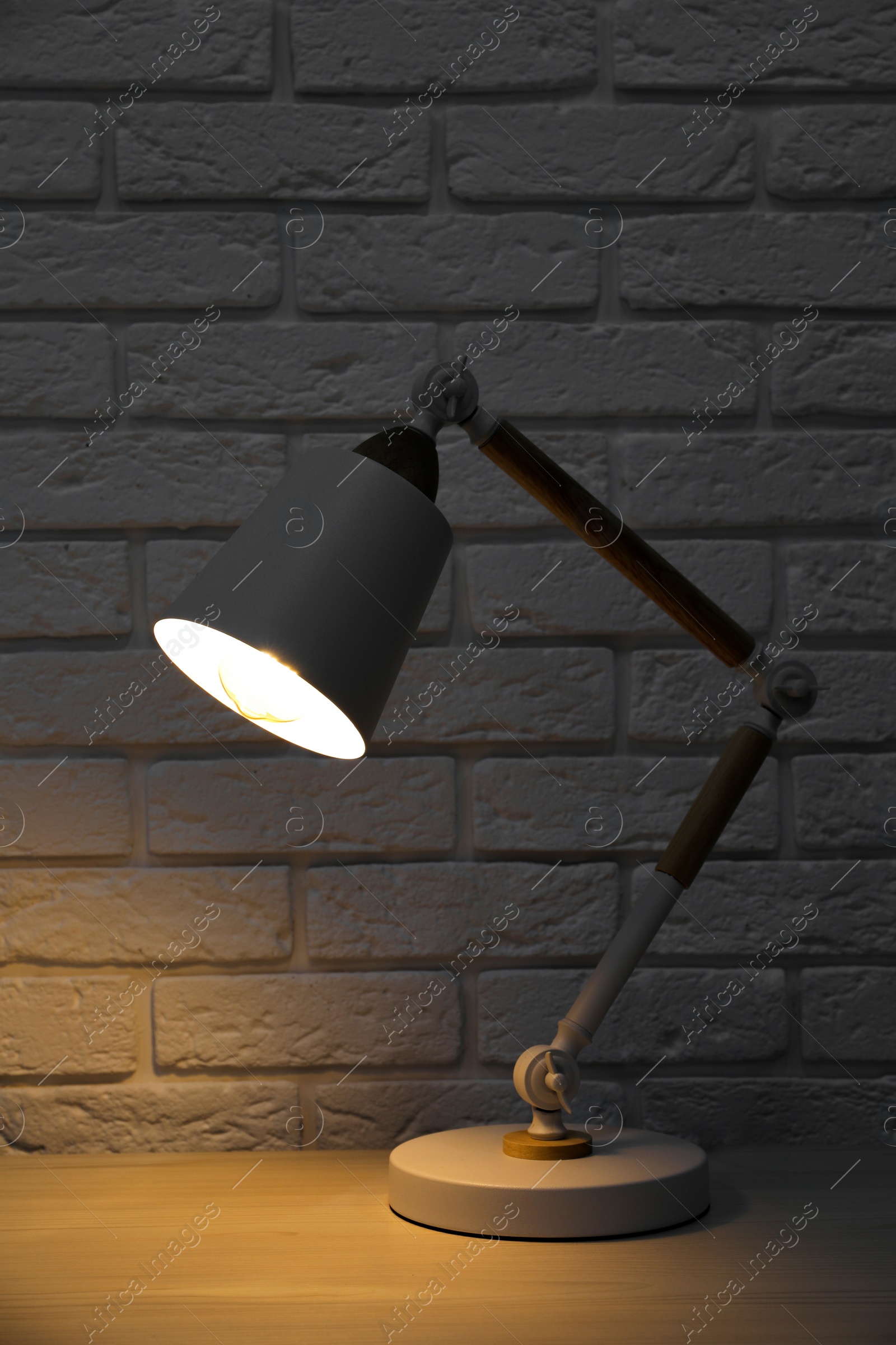 Photo of Stylish modern desk lamp on white wooden table near brick wall at night