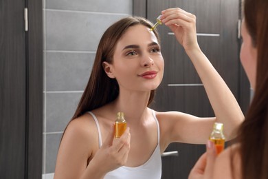 Photo of Young woman applying essential oil onto face in bathroom