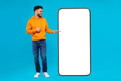 Image of Man pointing at huge mobile phone with empty screen on light blue background. Mockup for design