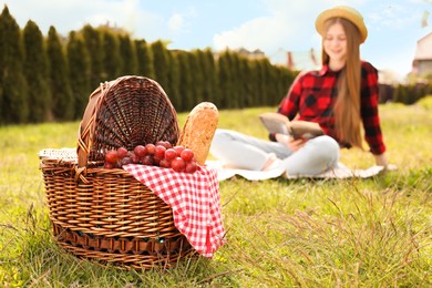 Photo of Girl reading book in park, focus on wicker picnic basket