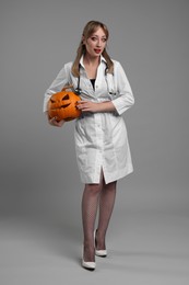 Photo of Woman in scary nurse costume with carved pumpkin on light grey background. Halloween celebration