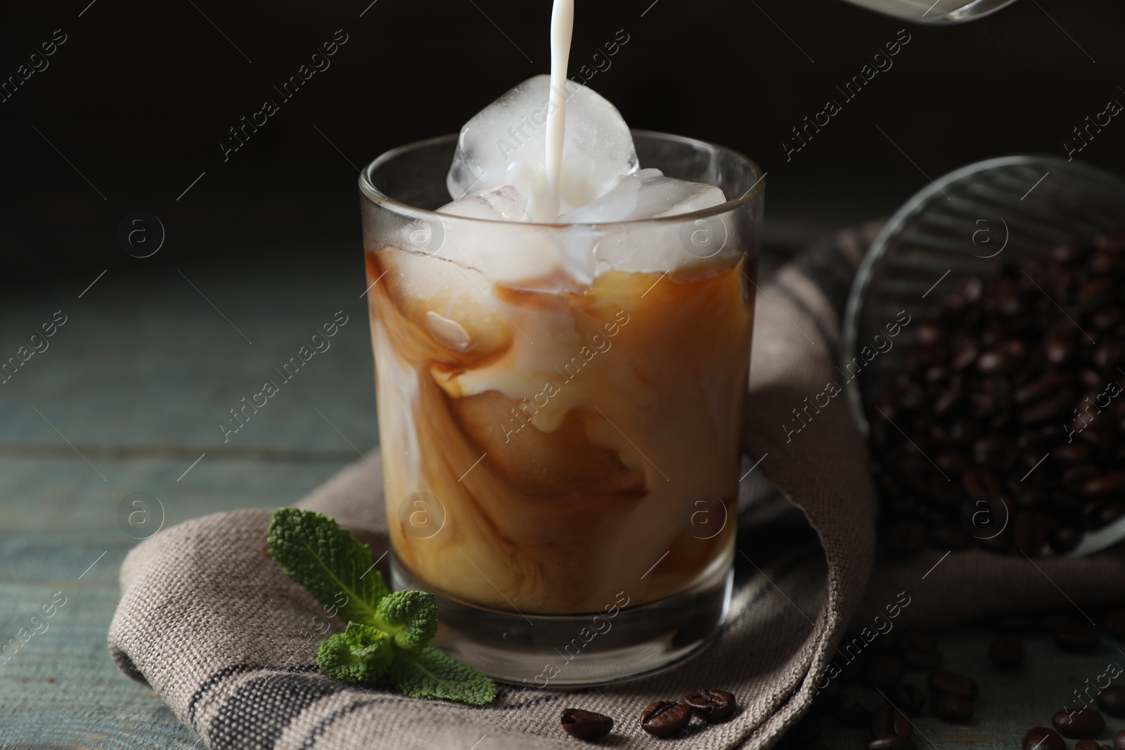 Photo of Pouring milk into glass of delicious iced coffee, beans and mint on light blue table, closeup
