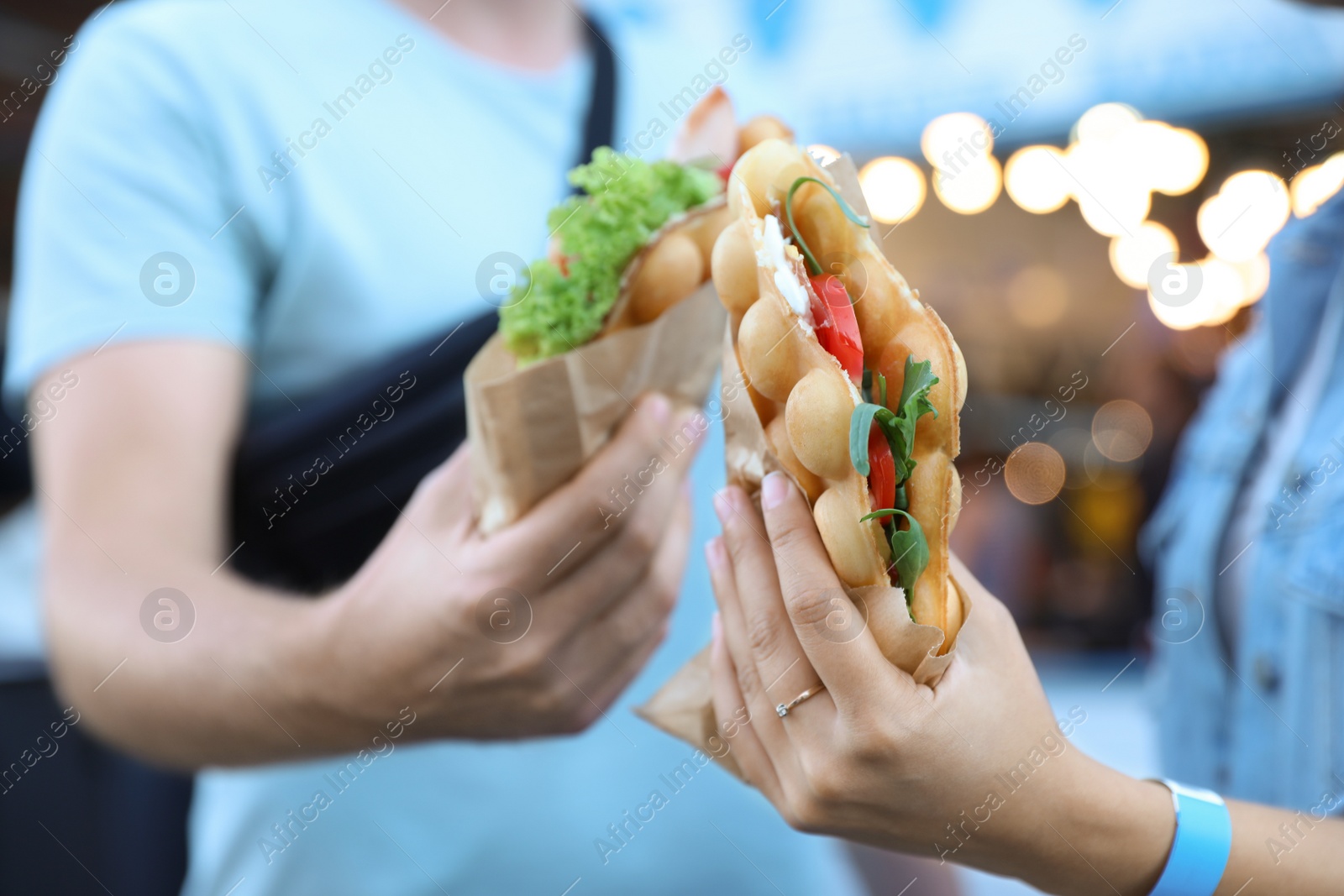 Photo of Young couple holding delicious bubble waffles with tomato and arugula outdoors, closeup