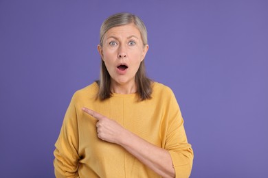 Surprised senior woman pointing at something on violet background