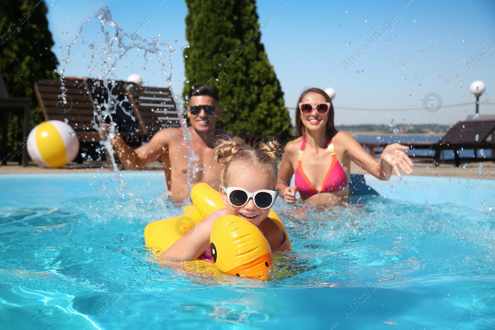 Photo of Little girl swimming with inflatable ring near her parents in outdoor pool on sunny summer day