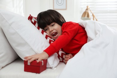Photo of Excited little boy finding gift box under pillow in bed at home. Saint Nicholas day tradition