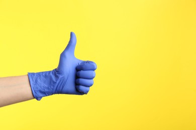 Photo of Person in medical gloves showing thumb up on yellow background, closeup of hand. Space for text