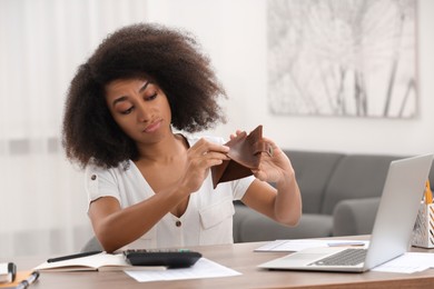 Confused woman with empty wallet planning budget at table in room. Debt problem