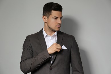 Man fixing handkerchief in breast pocket of his suit on light grey background