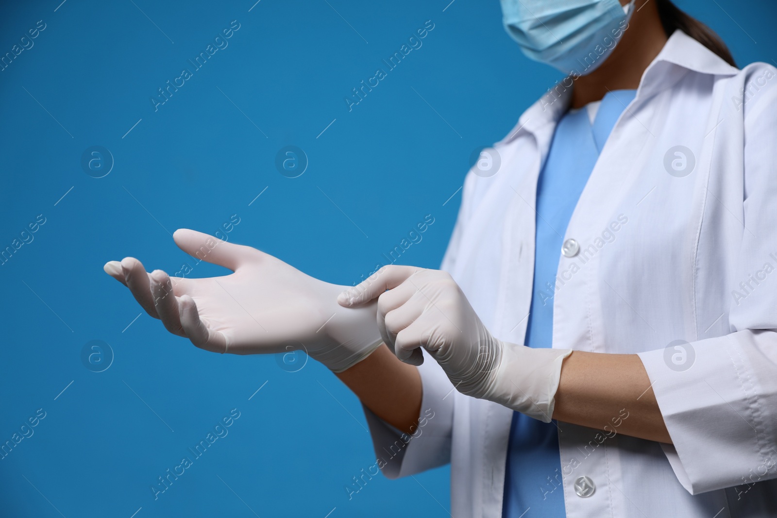 Photo of Doctor in protective mask putting on medical gloves against blue background, closeup