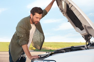 Upset man looking under hood of broken car outdoors