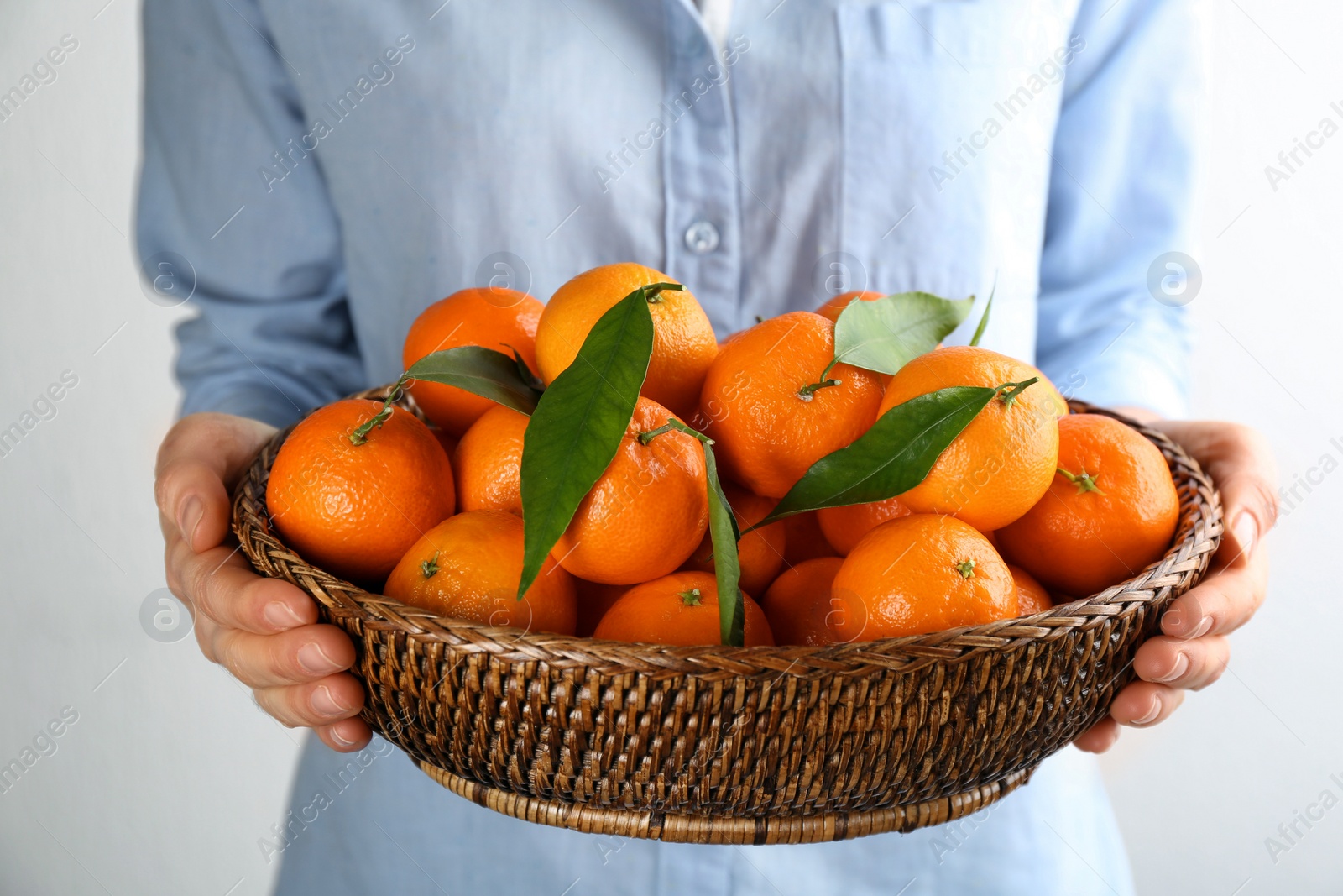 Photo of Woman holding wicker bowl with fresh tangerines on light background, closeup