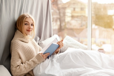 Beautiful young woman in knitted sweater sitting with book near window at home. Winter atmosphere