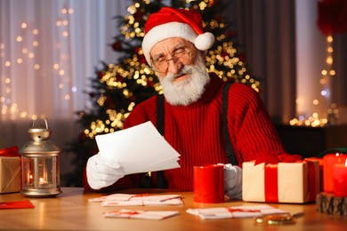 Photo of Santa Claus reading letter and drinking hot beverage at his workplace in room decorated for Christmas