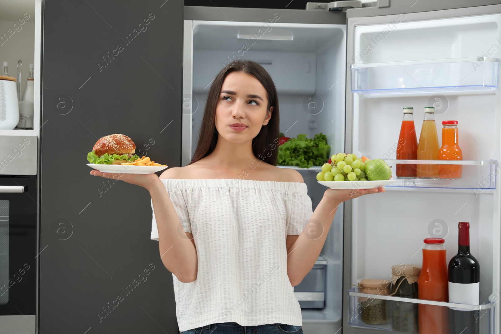 Photo of Woman choosing between fruits and burger with French fries near refrigerator in kitchen