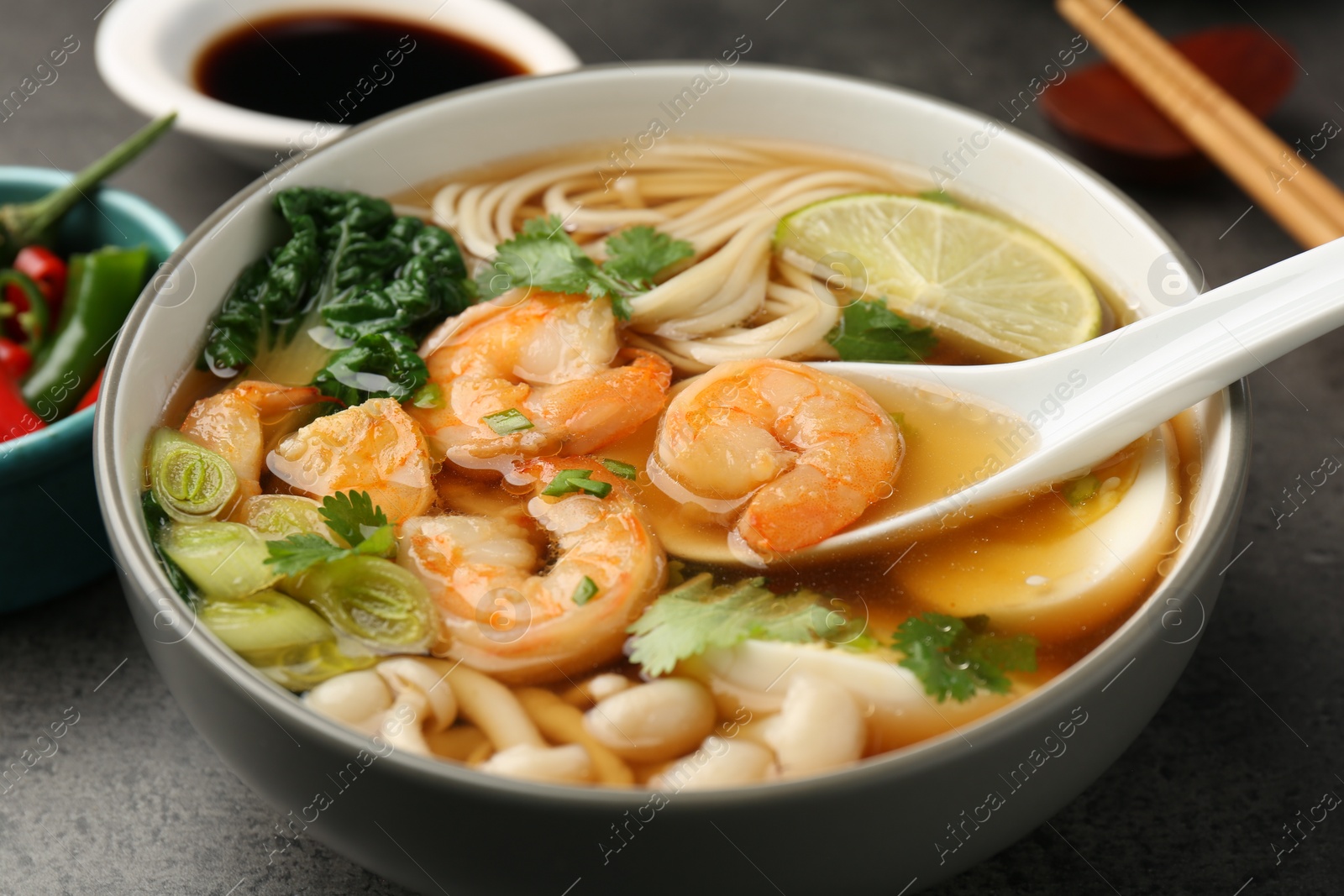 Photo of Eating delicious ramen with shrimps from bowl with spoon at grey table, closeup. Noodle soup