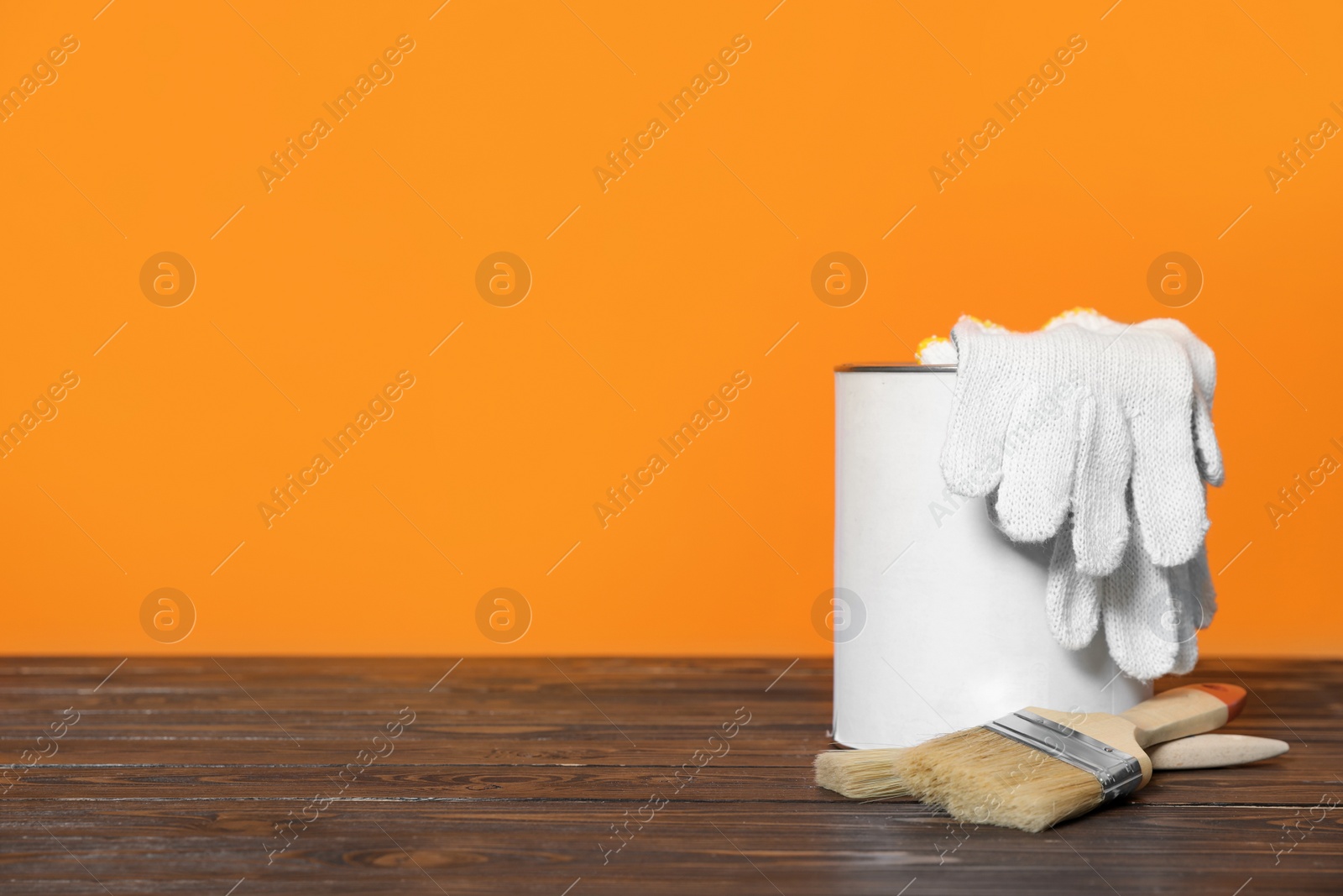 Photo of Can of paint, gloves and brushes on wooden table against orange background. Space for text
