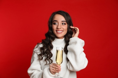 Beautiful young woman in warm sweater holding glass of champagne on red background. Christmas party