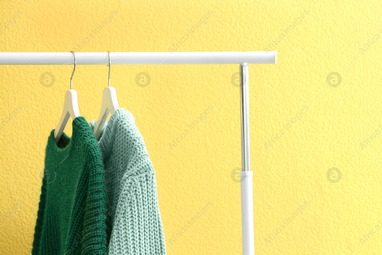 Photo of Warm sweaters hanging on rack near yellow wall, closeup