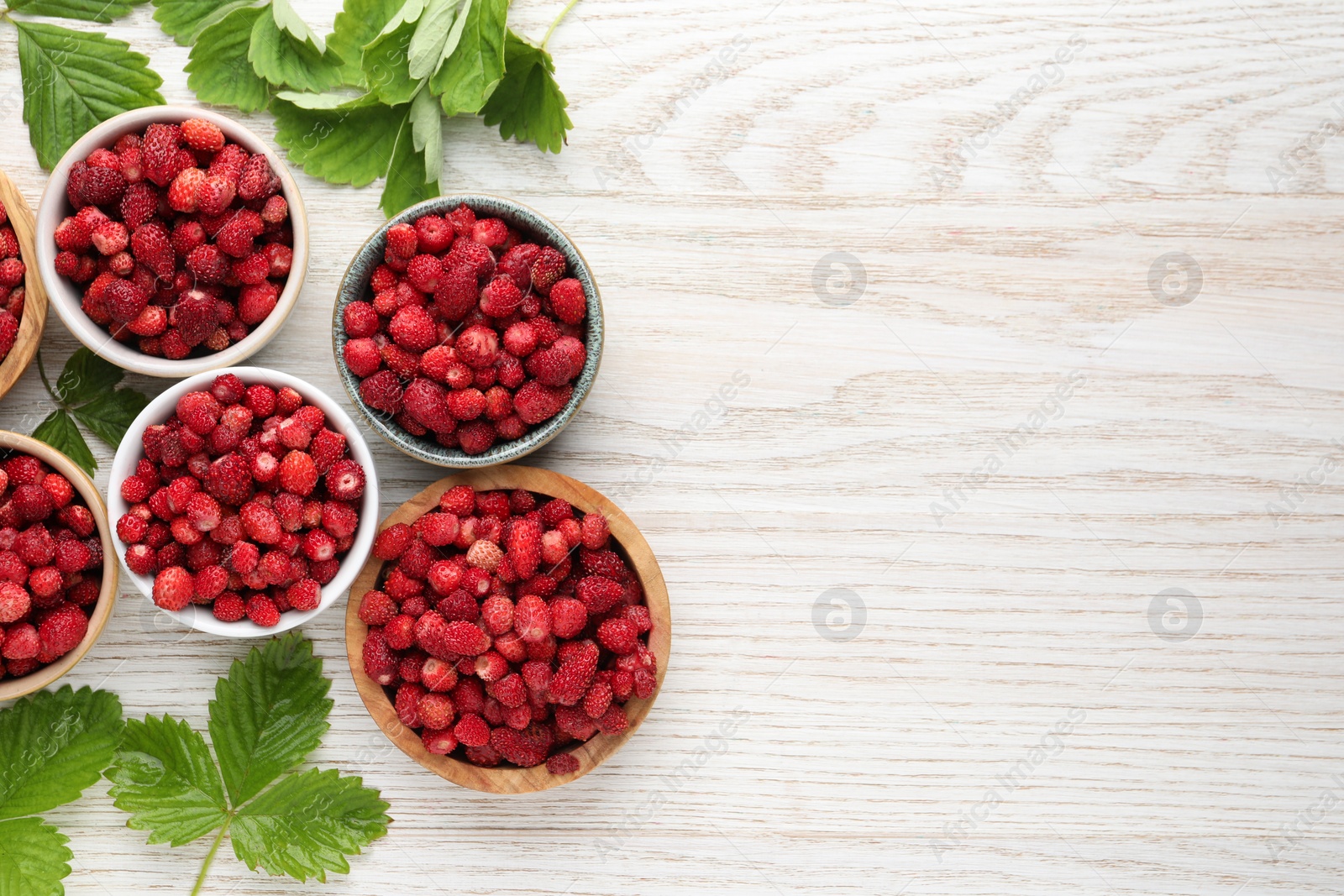 Photo of Fresh wild strawberries in bowls and leaves on white wooden table, flat lay. Space for text