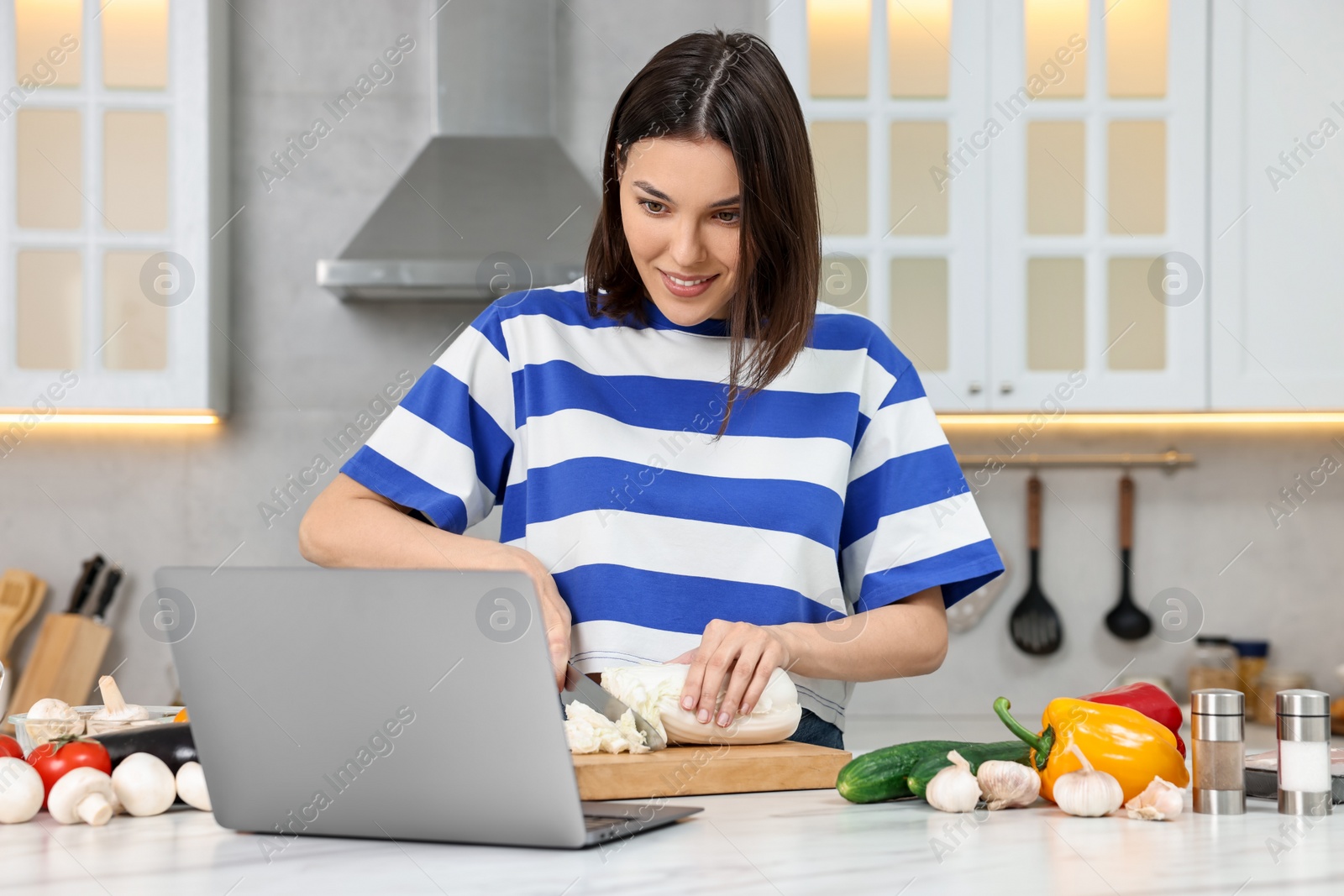 Photo of Young woman cooking while watching online course via laptop in kitchen