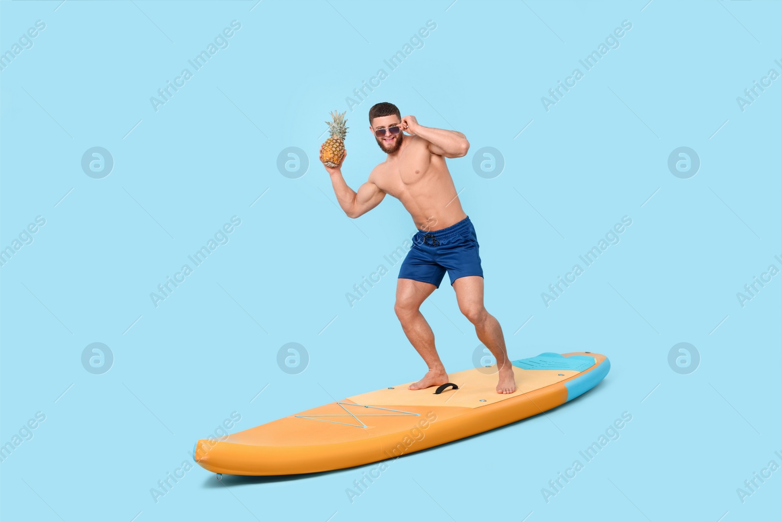 Photo of Happy man with pineapple posing on SUP board against light blue background