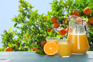 Image of Fresh orange juice on light blue wooden table in orchard. Space for text