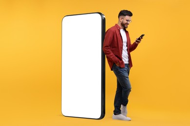 Image of Man with mobile phone standing near huge device with empty screen on orange background. Mockup for design