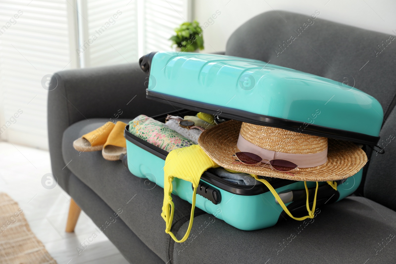 Photo of Open suitcase full of clothes, shoes and summer accessories on sofa in room