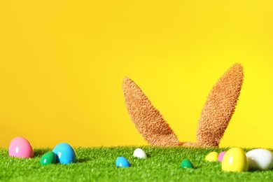 Easter eggs on green lawn and funny bunny ears against color background, space for text