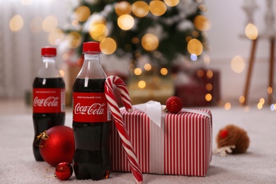 Photo of MYKOLAIV, UKRAINE - JANUARY 13, 2021: Bottles of Coca-Cola, candy cane, gift box and Christmas balls on floor in room