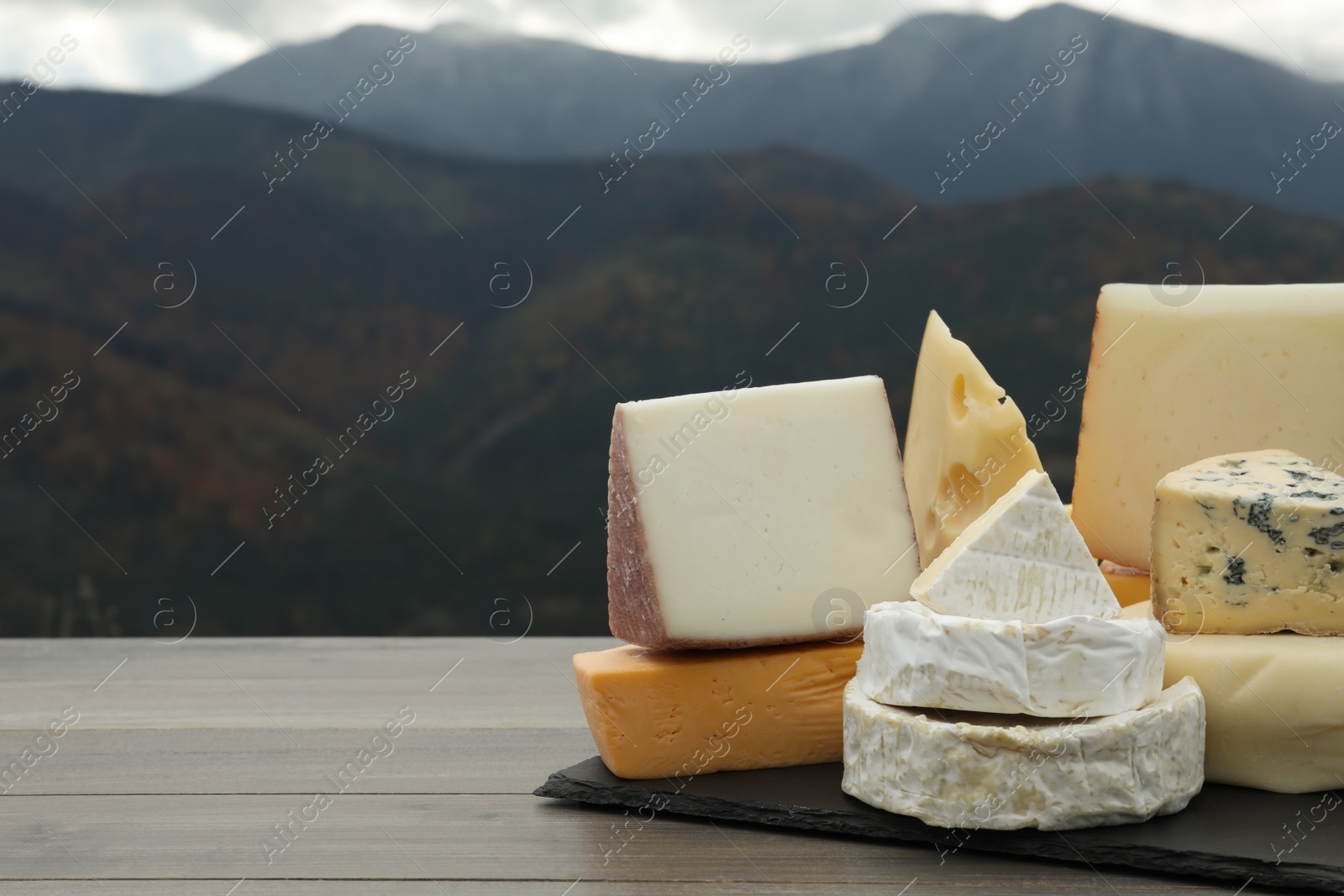 Photo of Different types of delicious cheeses on wooden table against mountain landscape. Space for text