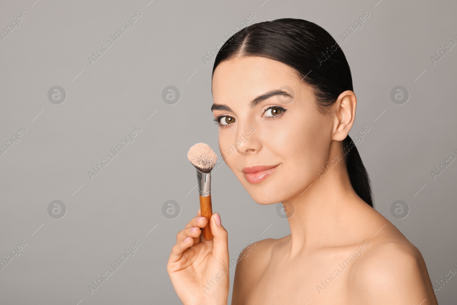 Photo of Beautiful young woman applying face powder with brush on grey background. Space for text