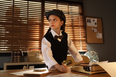 Photo of Cute little detective with magnifying glass at table in office