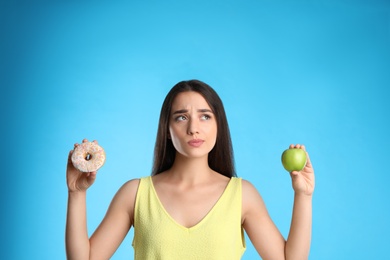 Photo of Doubtful woman choosing between apple and doughnut on light blue background